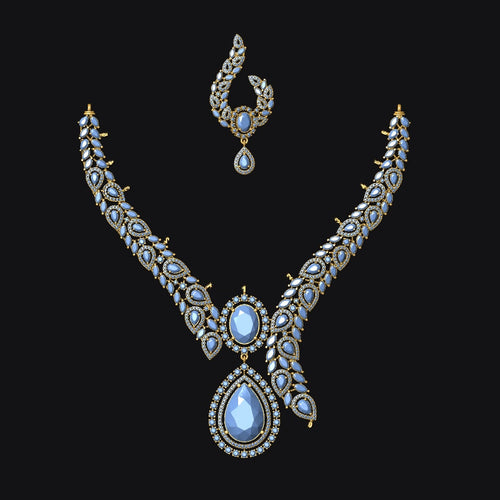 An ornate gold-toned jewelry set featuring a V-shaped necklace and a matching topkara earring, both adorned with small clear stones and a central teardrop-shaped light blue gemstone, set against a solid black background.