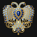 An ornate gold-toned brooch or pendant featuring a symmetrical winged design, set with deep blue teardrop-shaped stones and numerous small pale blue and white stones, shown on a solid black background.