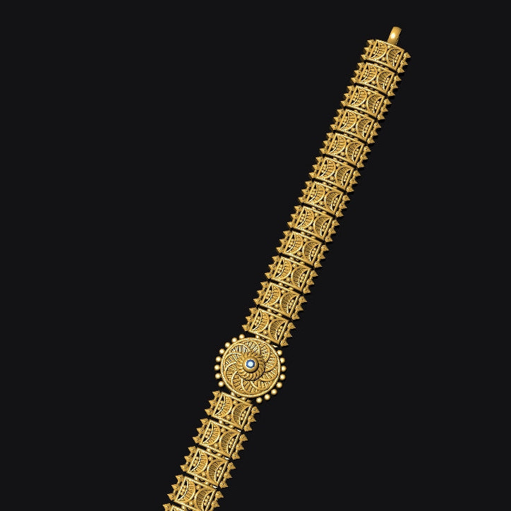 A gold-toned ornate bracelet or anklet with a cylindrical patterned body and a circular medallion detail, shown diagonally against a solid black background.