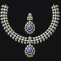 Gold-toned traditional Indian jewelry set featuring a necklace and matching pendant, embellished with white stones and blue teardrop gemstones, displayed on a solid black background.